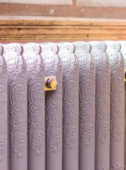 Detail of Rococo III Ornate cast Iron radiator finished in lilac with brass retaining stays by yellow easy chair and floral curtains