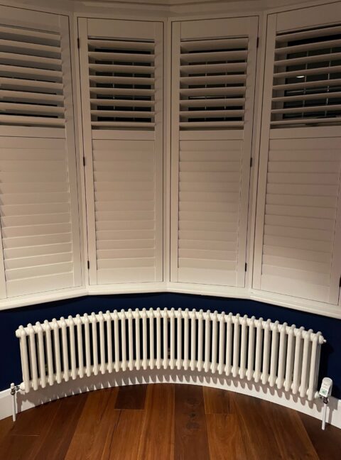 Long Bay Window Steel Column Tuba Radiator with white shutters and dark wooden floor