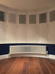 Long Curved bay wiondow radiator Tuba steel column finished in white with dark wooden floor and shutters