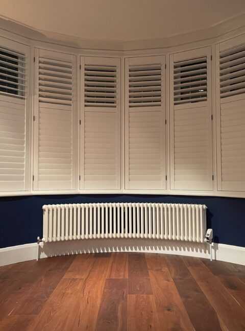 Long Curved bay wiondow radiator Tuba steel column finished in white with dark wooden floor and shutters